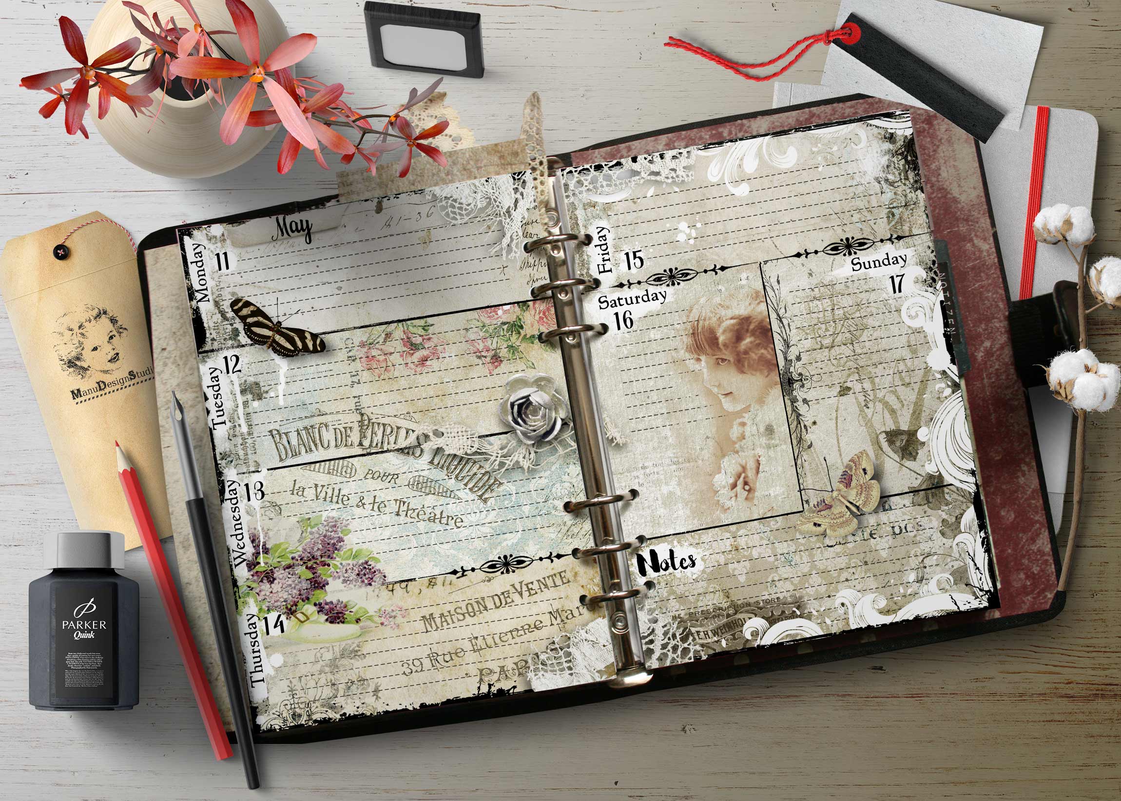 Open vintage-themed planner laid flat on a wooden desk, surrounded by red flowers, lace, tags, and craft supplies like ink and pencils.